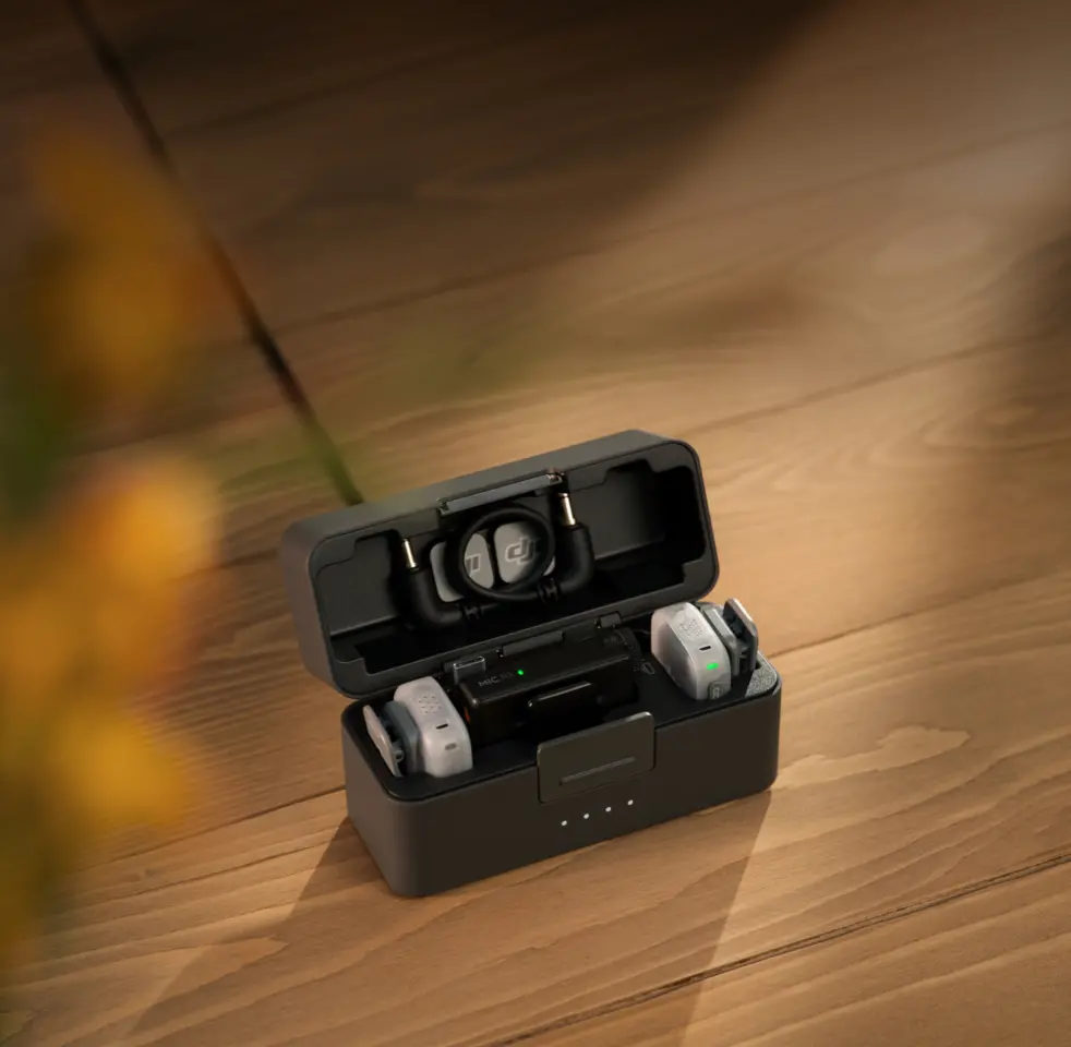 An open DJI wireless microphone charging case sits on a wooden surface, revealing the receiver and two transmitters inside along with a stored audio cable.